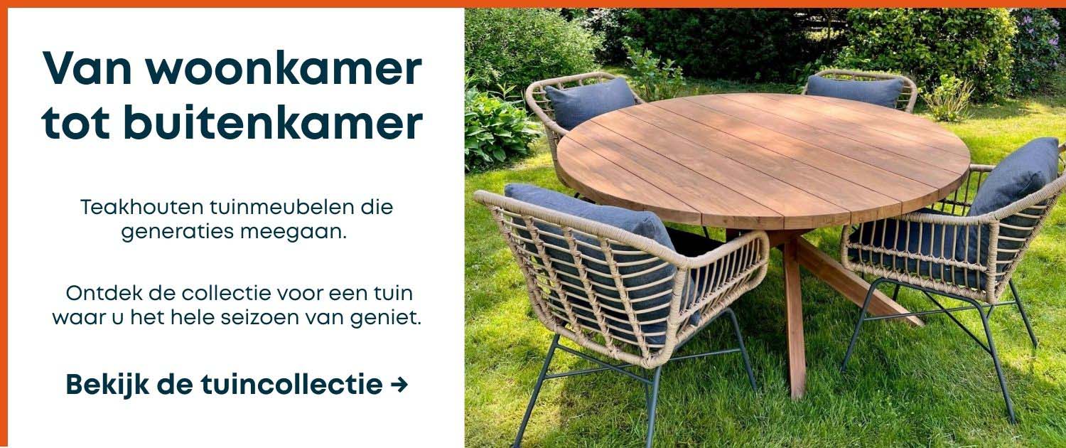 Ronde teakhouten tuintafel met vier gevlochten stoelen met donkergrijze kussens, opgesteld op een groen gazon omringd door weelderige tuinbeplanting